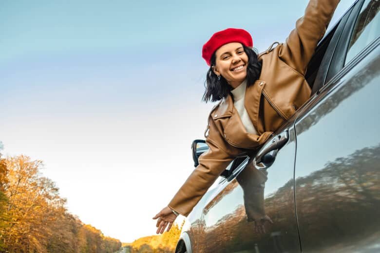 a smiling woman in a red beret looks out of the car window on the roadside in autumn